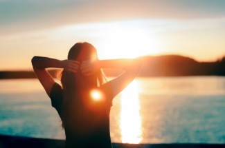 Frau hält Arme hinter ihrem Kopf und steht am Meer im Sonnenuntergang