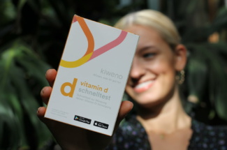 Frau hält Vitamin D test von kiweno in die Kamera vor Pflanzen im Hintergrund