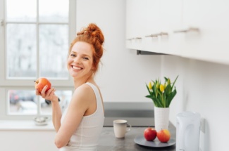 Frau steht lächelnd in Küche und hält einen Apfel in der Hand
